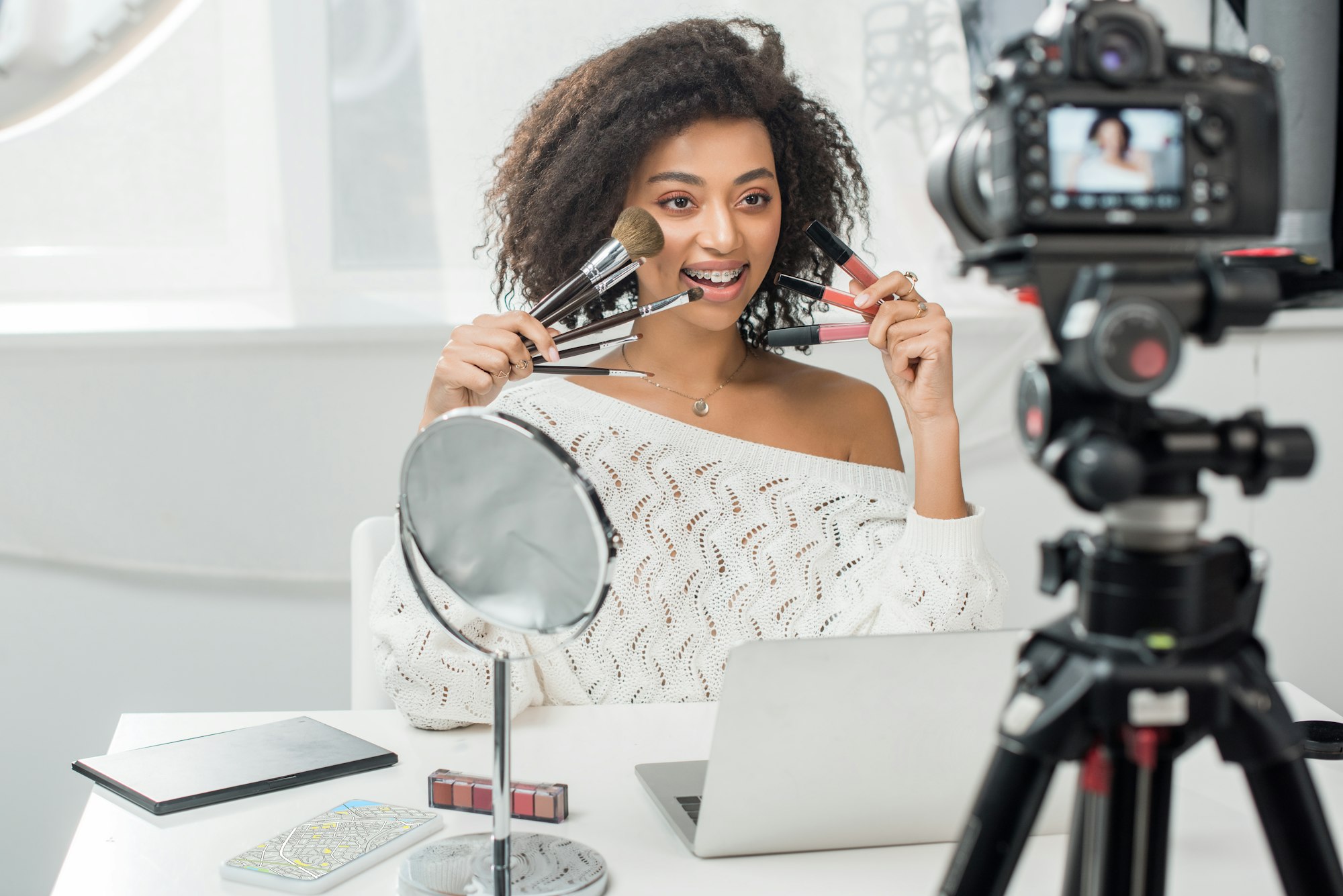 selective focus of happy african american influencer in braces holding lip glosses and cosmetic